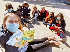A teacher takes a selfie style photo holding two books sitting on the sidewalk with a half circle of students . 
