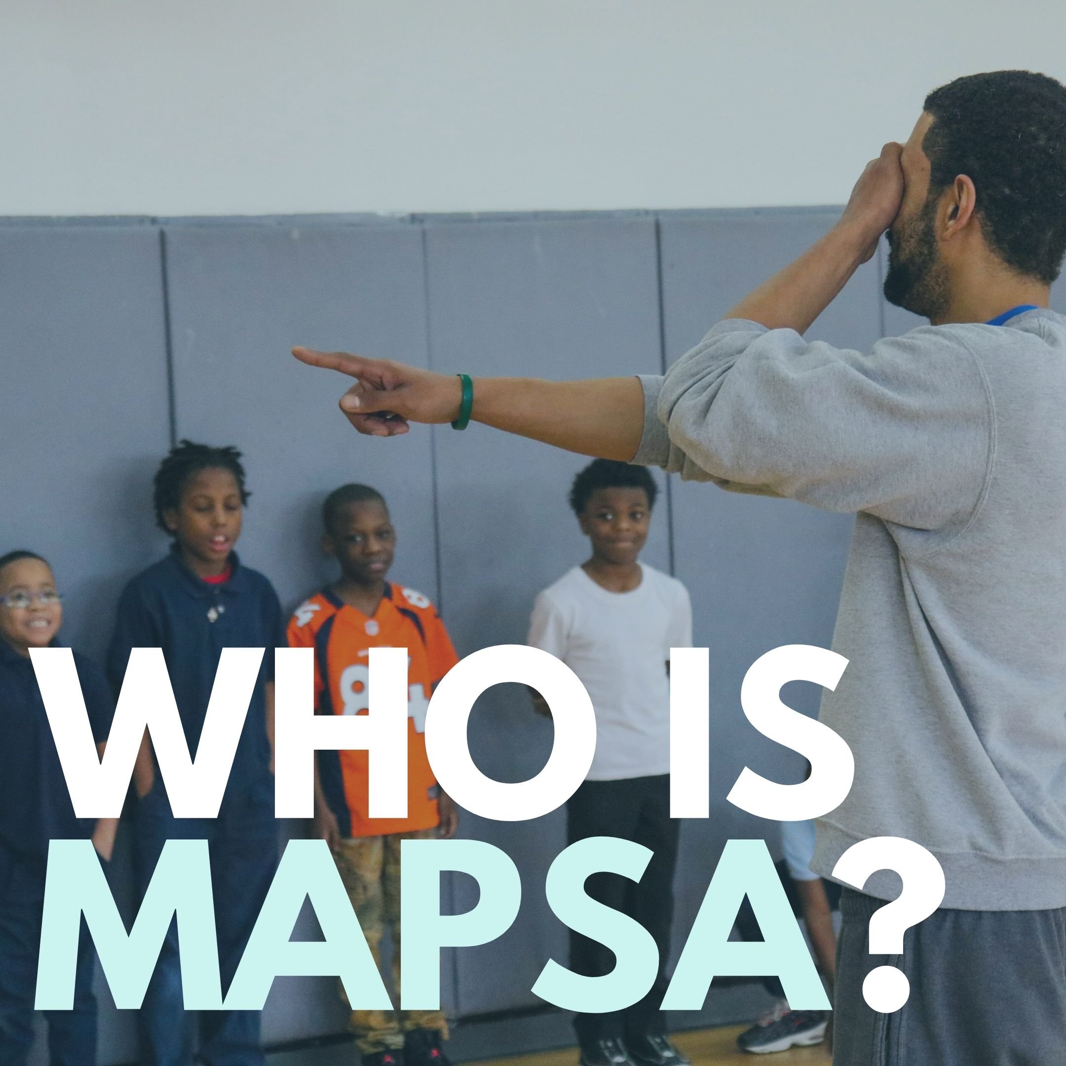 A photo of a black male gym teacher covering his eyes with one hand and with the other randomly selecting students for a game, who are lined up against one gym wall.