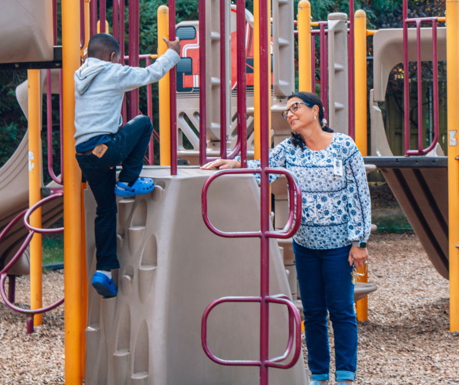 An African-American child playing on a playground with a teacher nearby watching him