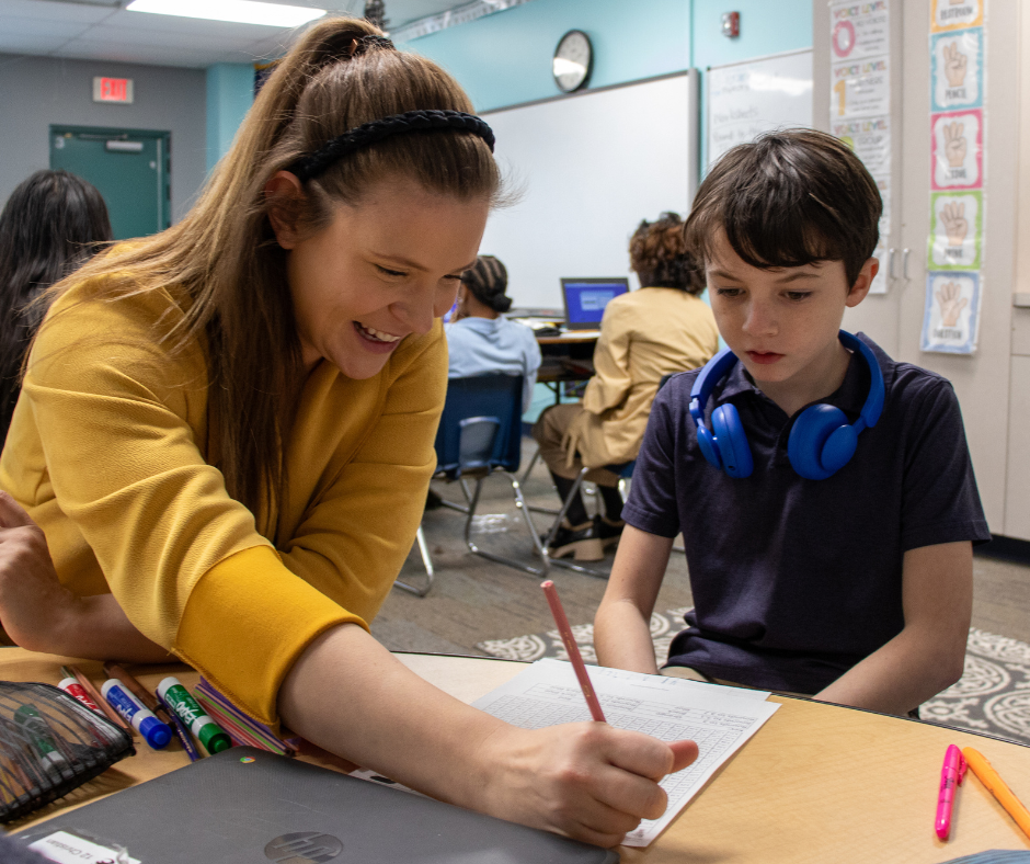 A white female teacher helping a young white boy with this school work