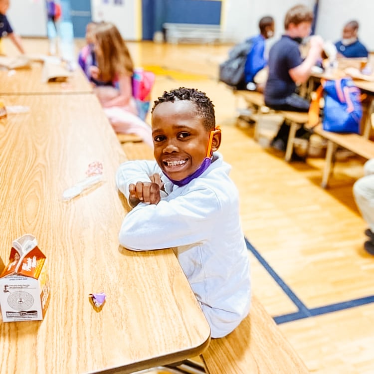 A photo of an elementary-aged, Black male student smiling with elbows resting on a cafeteria table.