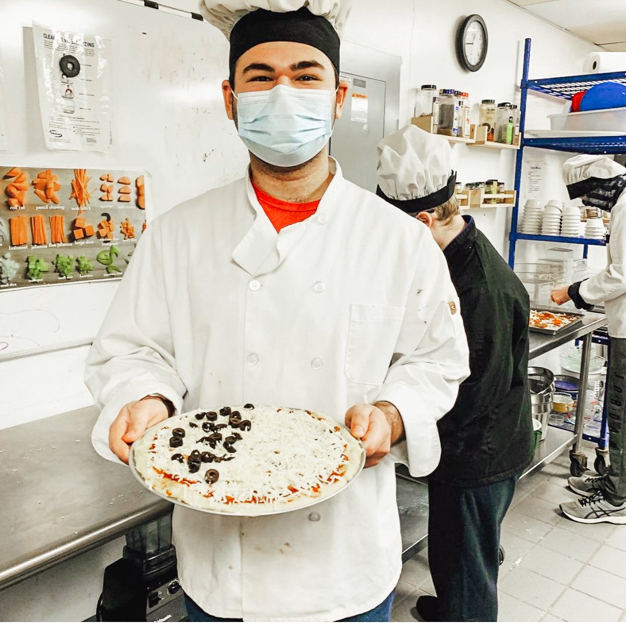 A white male student stands in a chefs coat and hat holding a pizza. 