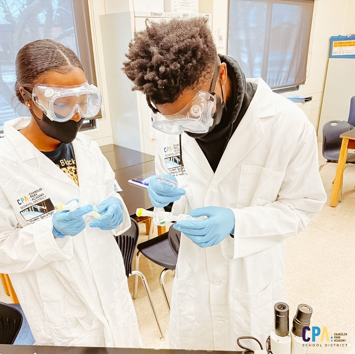 One female and one male African American students stand over their science experiment wearing lab coats, goggles, and gloves 