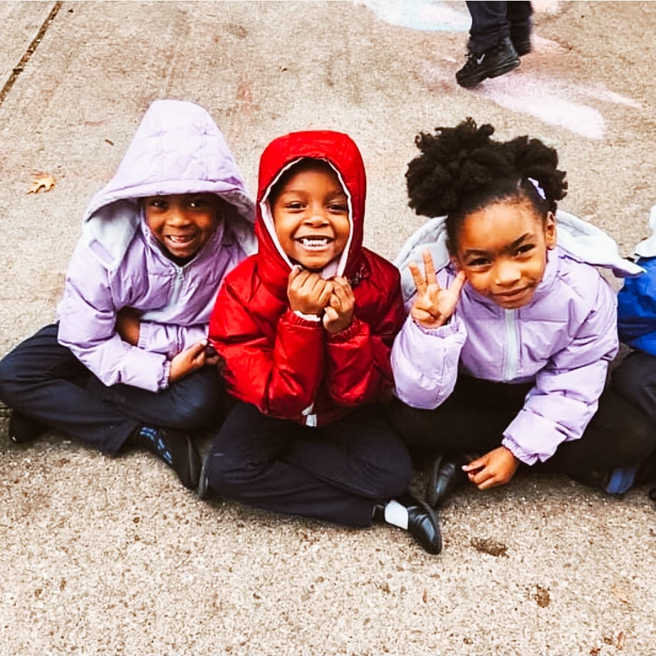 Two African American female students in purple winter jackets sit on the sidewalk on either side of a male African American student wearing a read jacket and smiling at the camera