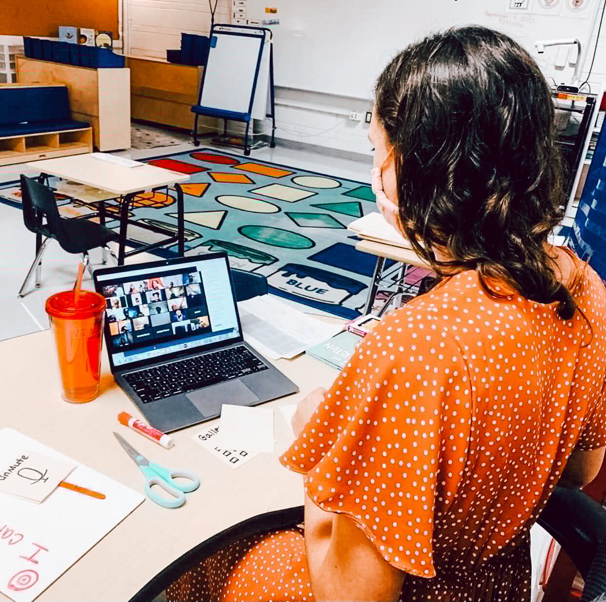 A female teacher wearing an orange dress sits at a desk talking to students on a Zoom video conference