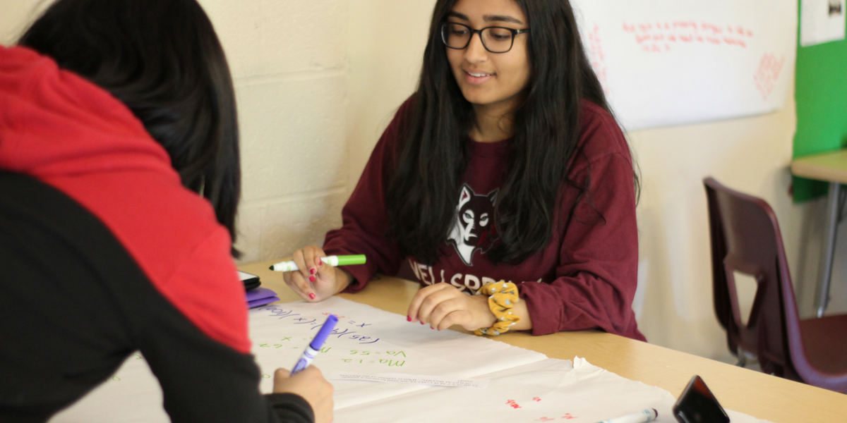 A photo of two female students practicing math equations at their desks using white paper and markers.
