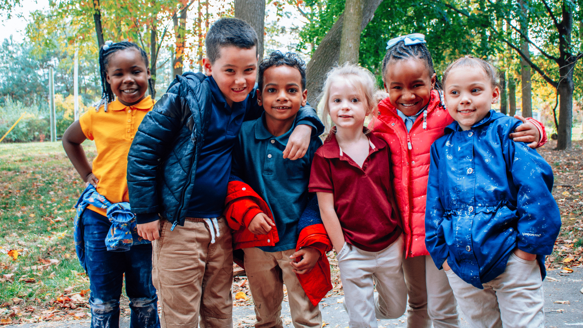 A group of 6 young children wearing bright colors standing outside with their arms around each other.
