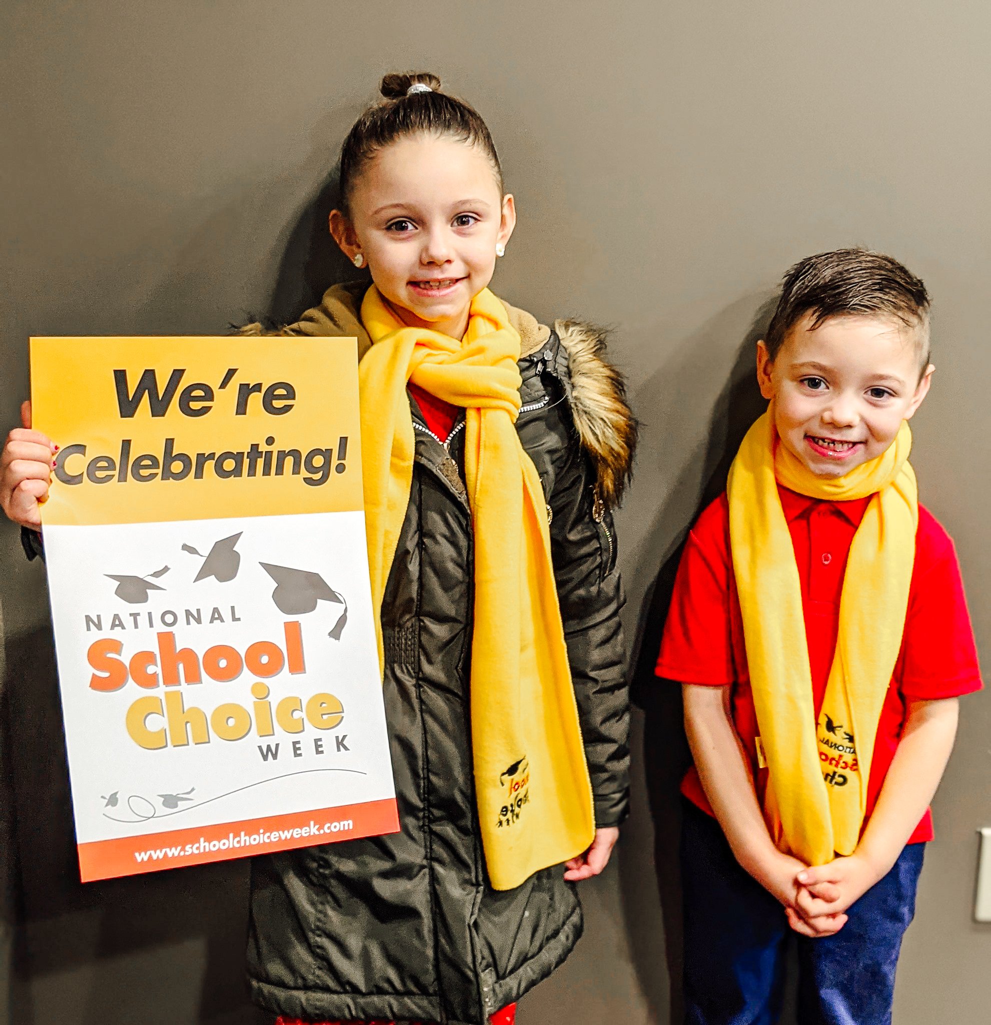 Two white students, a taller female and a male on the right smile wearing yellow National School Choice Week scarfs. The female student holds a sign reading "We're Celebrating! National School Choice Week" 