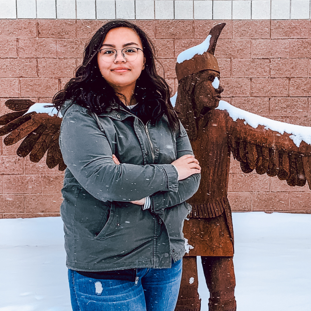 A photo of a high-school aged, Native American female student, posing in front of a Native American statue.