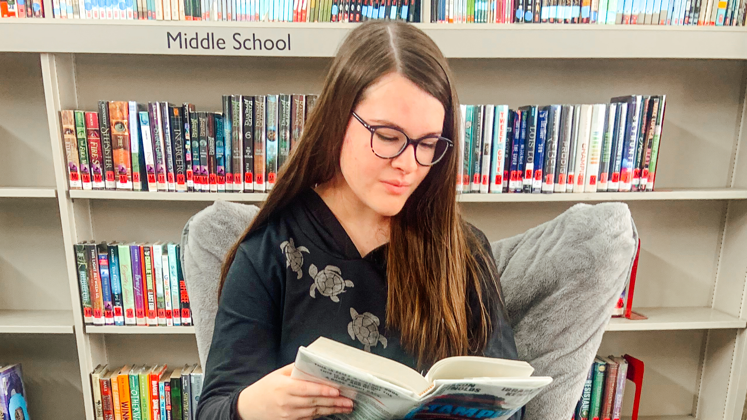 A photo of Kingsbury Country Day School 8th grader Ruby Howe sitting in a chair in the library reading a book. 