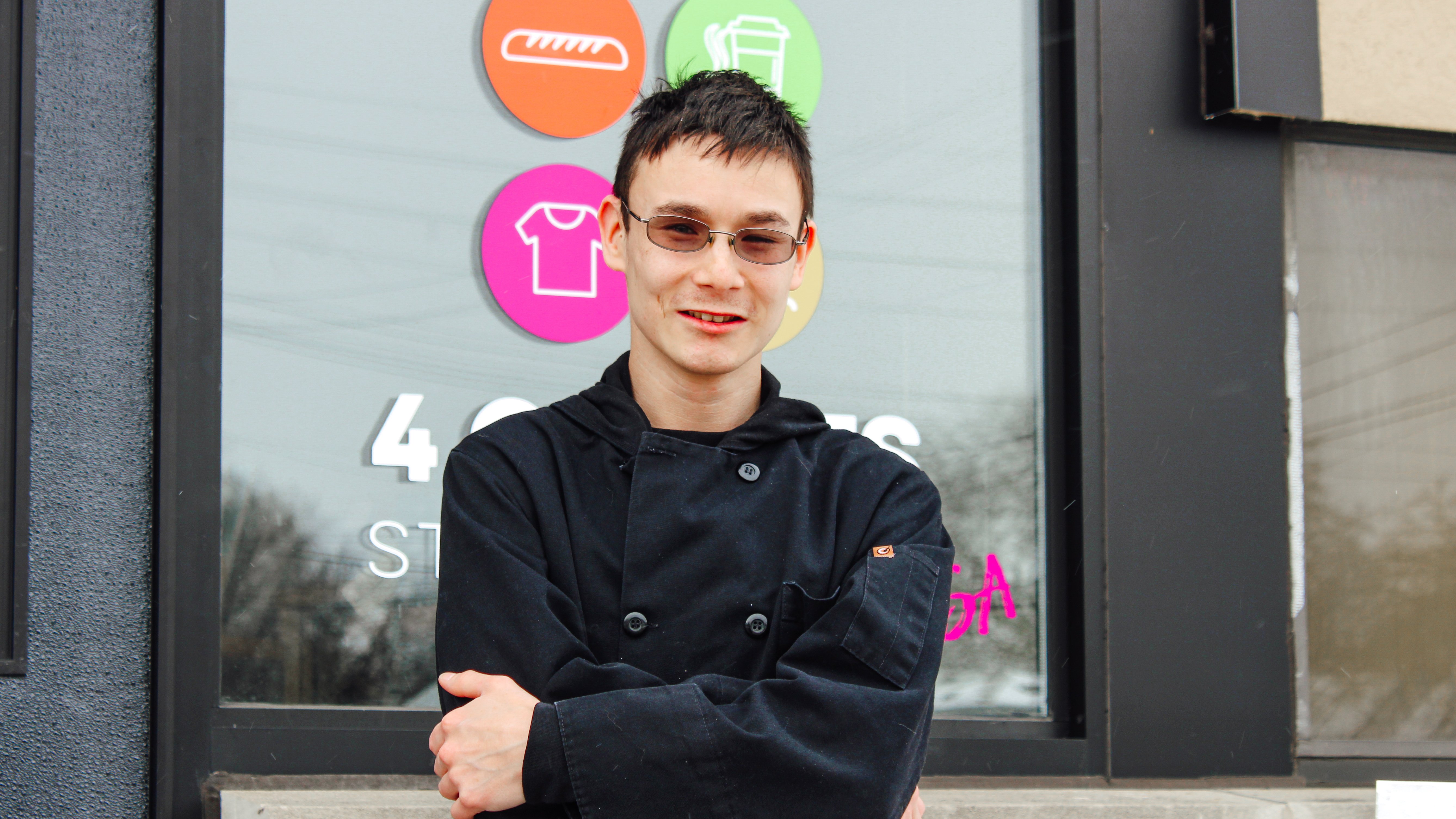 Rising Stars Alumni Josh Kowalski stands with his arms crossed and smiling in front of the bakery store front used by Rising Star students. 