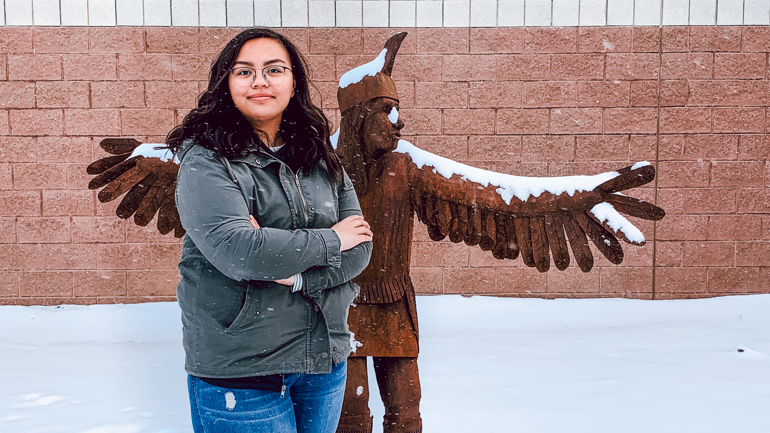 A photo of Josephine Troxell, high school senior at Nah Tah Wahsh Public School Academy, standing in front of a Native American wooden sculpture on her school grounds.