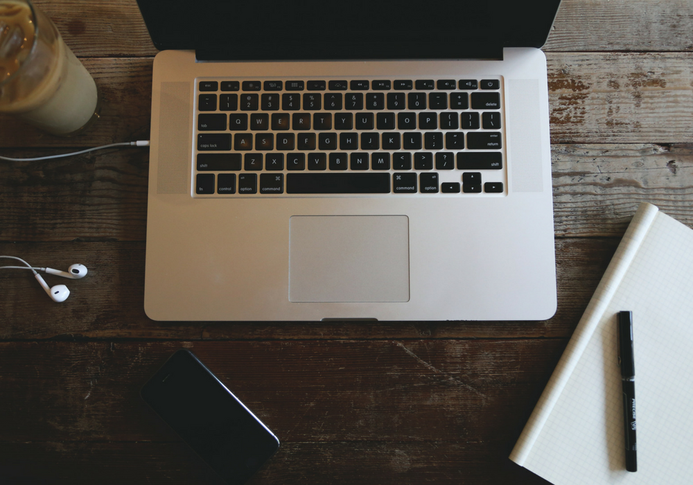 A photo of a laptop next to a notebook, pen and headphones.