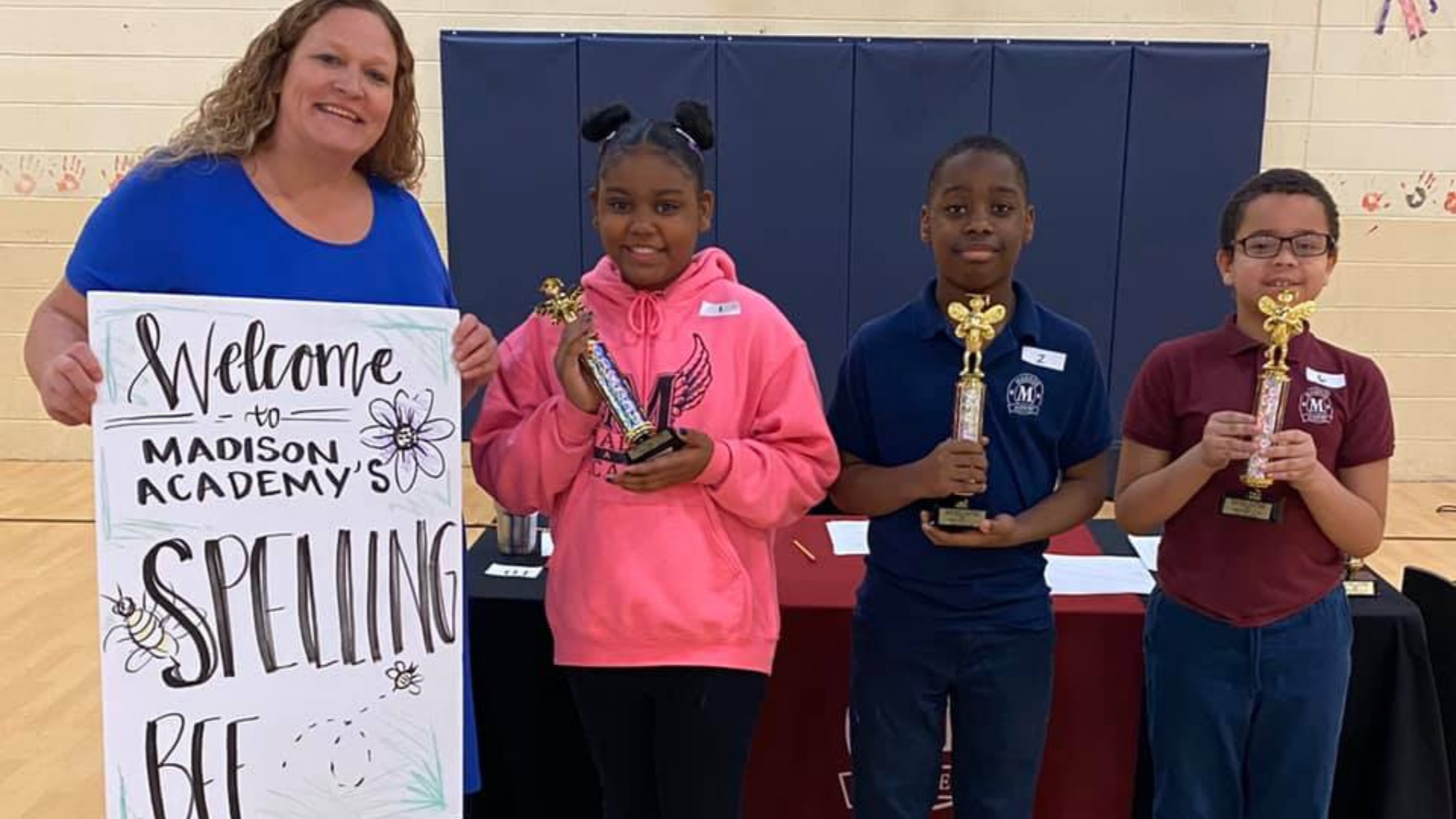 A white teacher stands holding a sign that reads welcome the Madison Academy's Spelling Bee