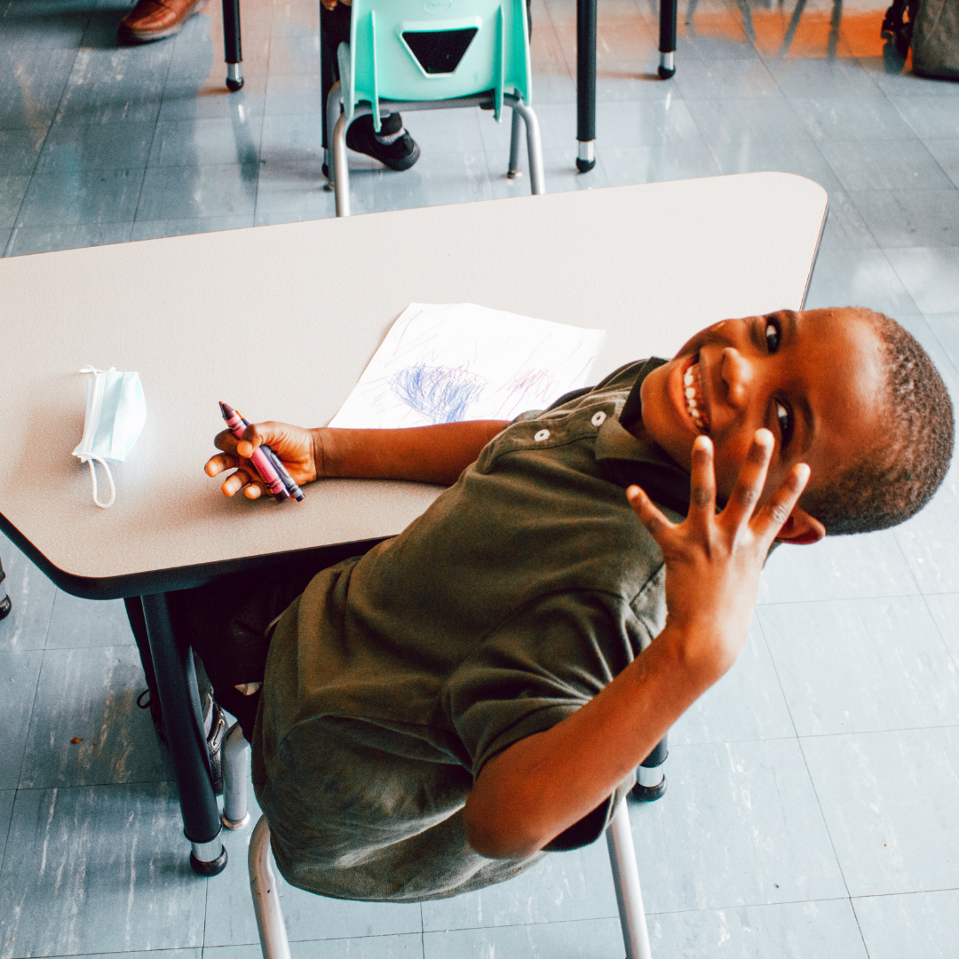 A photo of an African American male student from Barack Obama Leadership Academy, seated at his desk and leaning back to look back at the camera, holding up a peace sign with is left hand, smiling.
